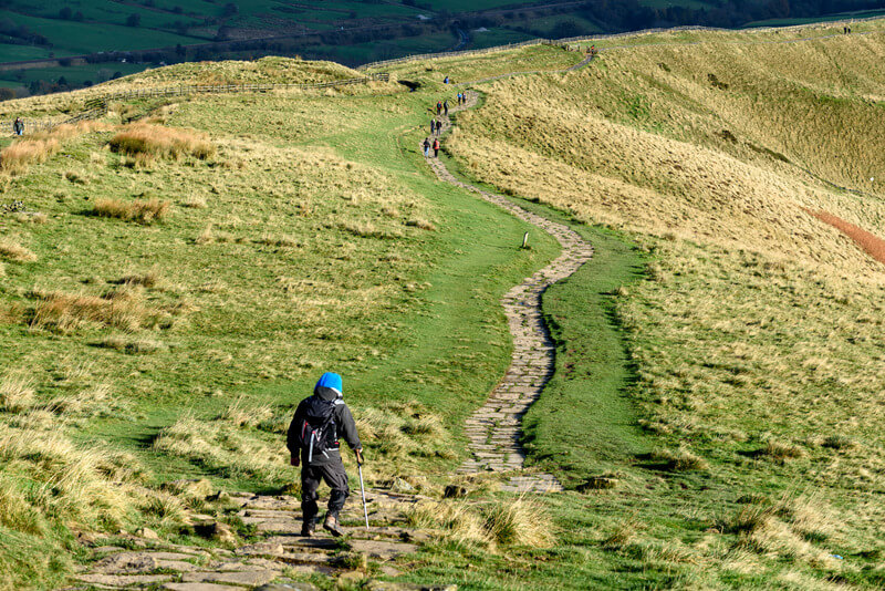 Walking trail in the lush and grassy Peak District, England. There is a man in the foreground and a winding trail is visible.