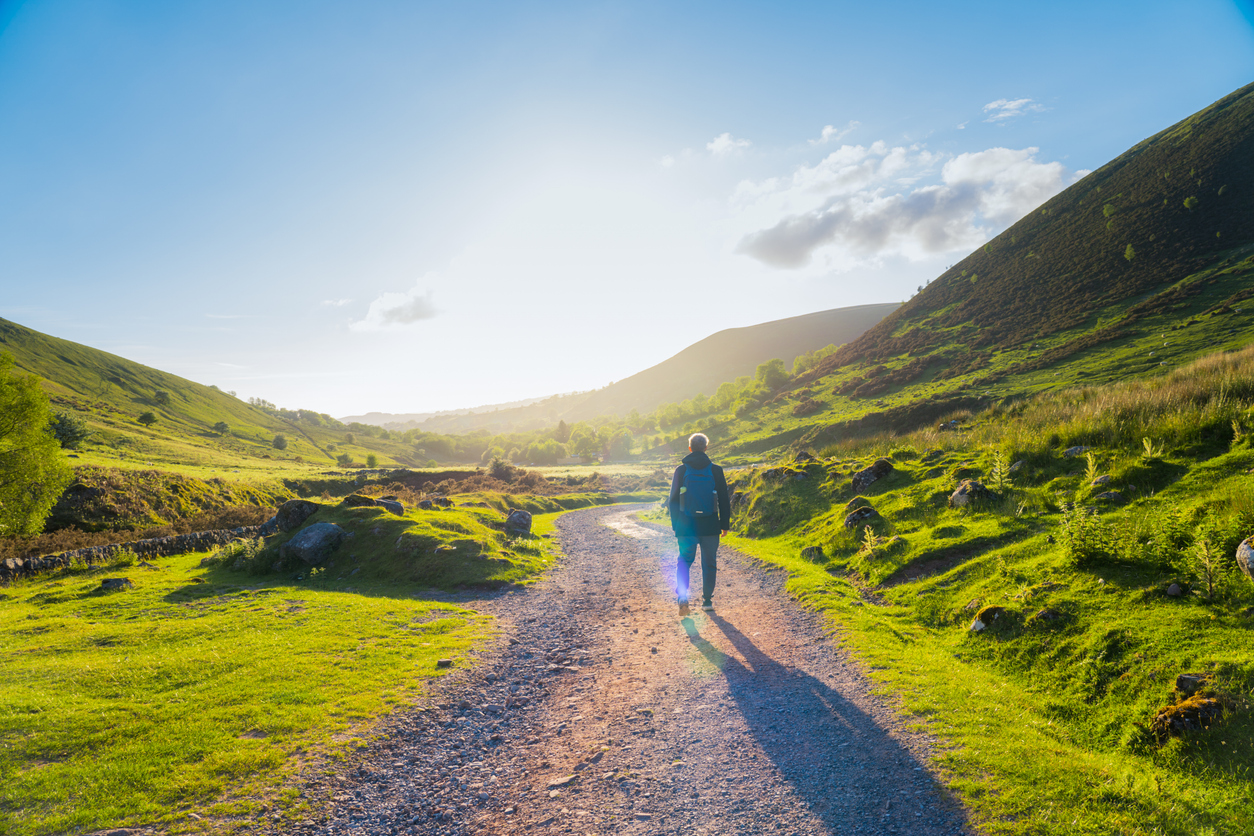 Image of a tourist with a backpack walking in North Wales on a mountain road with green hills around. Hiking alone. Sunset light. Wales, Snowdonia