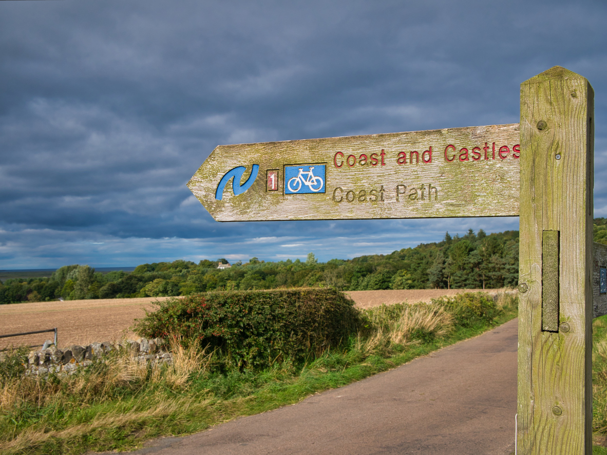 This way to your next coastal caravan break. A wooden sign post points the way of the Coast and Castles cycle route and Northumberland Coast Path. Taken in sunshine against a cloudy sky in autumn.