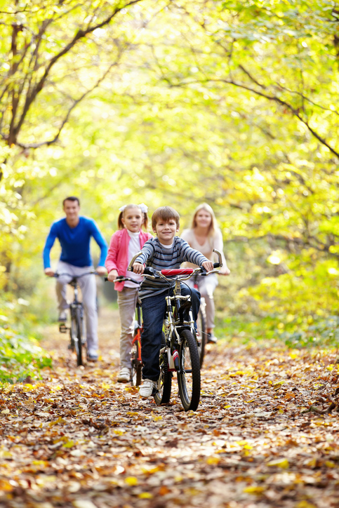 This image depicts a joyful family cycling together on a woodland trail during autumn. In the foreground, a young boy leads the way on his bike, beaming with excitement. He is followed by a little girl and two adults, possibly his sister and parents, all on bicycles. The path is blanketed with fallen leaves, reflecting the vibrant, golden hues of the trees around them. The scene captures a moment of active, familial happiness surrounded by the natural beauty of the season.