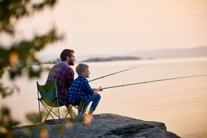 Side view portrait of father and son sitting together on rocks fishing with rods in calm lake waters with landscape of setting sun, both wearing checkered shirts, shot from behind tree