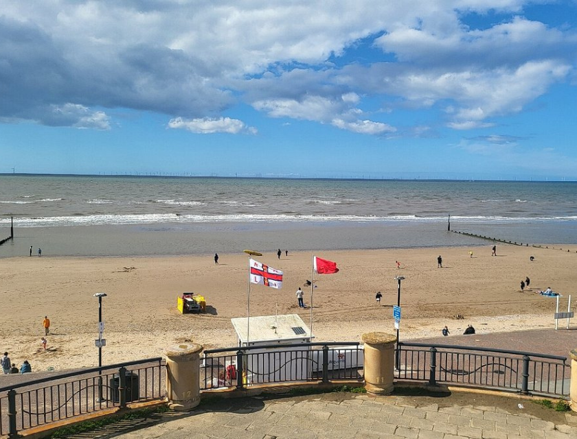 The image shows a coastal scene at a beach in Towyn with a sandy shore and ocean under a partly cloudy sky. People are walking and relaxing on the beach, and two flags are prominently displayed near the center. A yellow vehicle is visible on the sand. The foreground features a railing and a paved area. Dog-Friendly Pubs Towyn are nearby for visitors to enjoy with their pets.