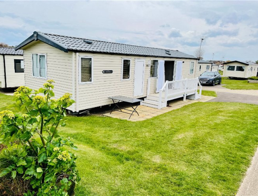 A light beige mobile home with a black roof and white trim is set on a well-maintained green lawn. The front of the home features a small white deck with railings and steps leading down to a paved patio area. A folding table is placed on the patio. In the foreground, there are green plants with yellowish flowers. Other mobile homes and a car are visible in the background under a cloudy sky. The overall scene is bright and well-kept, suggesting a pleasant holiday park environment.




