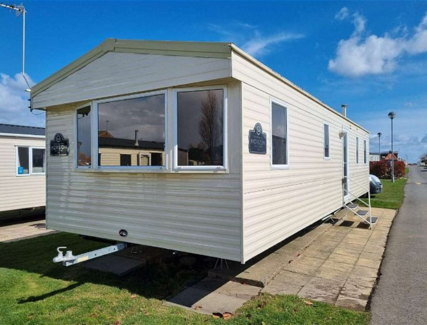 A light beige mobile home with large windows and white trim, positioned on a paved and grassy area at Primrose Valley Caravan Park. The home features a small set of metal steps leading to the entrance. The bright and clear blue sky enhances the inviting atmosphere. Other mobile homes and a road are visible in the background, indicating a well-organized and maintained park setting. This accommodation option is available through UK Caravans 4 Hire, providing a comfortable and convenient stay in a picturesque environment.