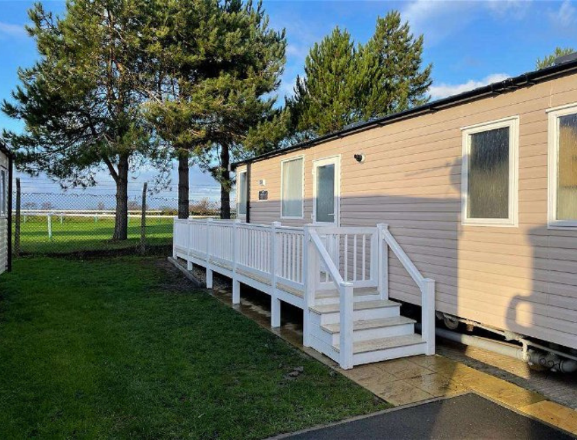 A static caravan with a white ramp and steps, situated in Pinewoods Caravan Holiday Park in Norfolk. The caravan is set against a backdrop of tall pine trees and a grassy area, offering a serene environment. This location provides convenient access to the picturesque walking trails in Norfolk, ideal for exploring the natural landscape.