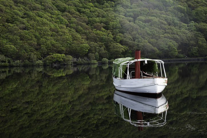 A lake tour boat sits on the glass-smooth water of Llyn Gwynant, which perfectly reflects both the boat and the Snowdon Mountain behind it. A wisp of cloud scuds along the trees behind the boat.