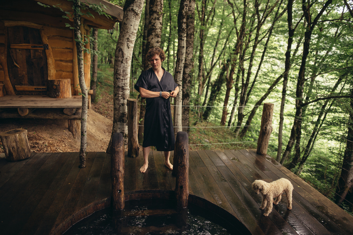 pleasant holidaymaker is taking off the black gown near the wooden bathtub and a dog. vacation and day off concept