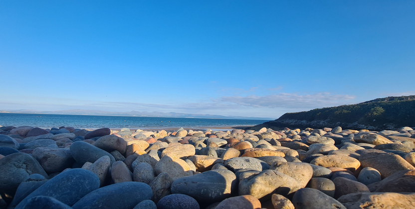 A scenic view of a pebble beach under a clear blue sky. The foreground is filled with smooth, multicolored pebbles of varying sizes, extending towards the calm ocean in the background. A grassy hill slopes down to the right, adding to the natural beauty of the scene. The distant horizon features a faint outline of mountains or distant landmasses across the water. This picturesque coastal landscape captures the serene and unspoiled nature of the beach.