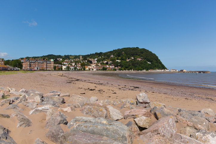 Image shows a coastal view of a sandy beach with scattered rocks in the foreground, under a clear blue sky. The sea is slightly visible. In the background, a quaint town town by the sea with brick buildings and houses nestled against a lush green hillside. The shoreline gently curves along the water's edge, with the ocean calm and inviting.