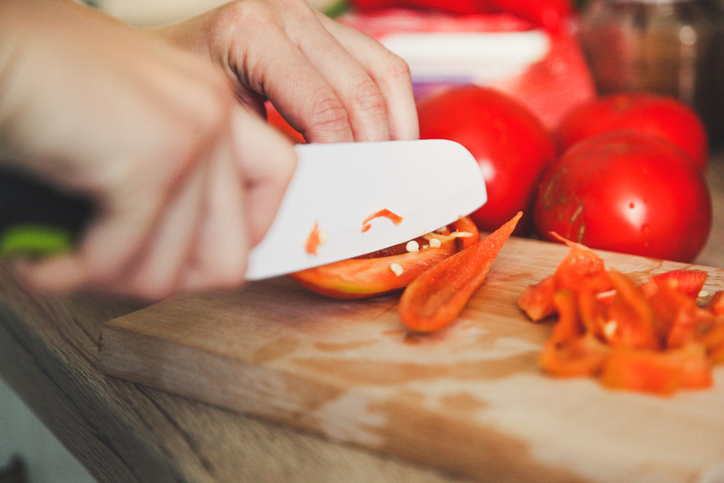 close up of a white knife slicing a red pepper with tomatoes in the background