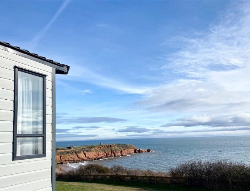 Image showing modern white cabin with a large window on the left, overlooking a scenic coastal landscape. The view features a lush green lawn that leads to cliffs made of red rock, extending into a calm blue sea. The sky is partly cloudy, suggesting a peaceful, bright day.