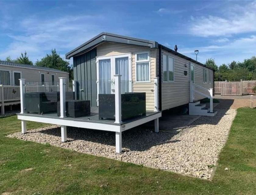 A beige mobile home with white trim and a slanted roof is positioned on a gravel area with a grassy lawn surrounding it. The front of the home features a large deck with glass railings and black outdoor furniture. Steps with white railings lead up to both the deck and the side entrance of the home. Another mobile home is visible to the left, and a wooden fence and more greenery are seen in the background under a clear blue sky.






