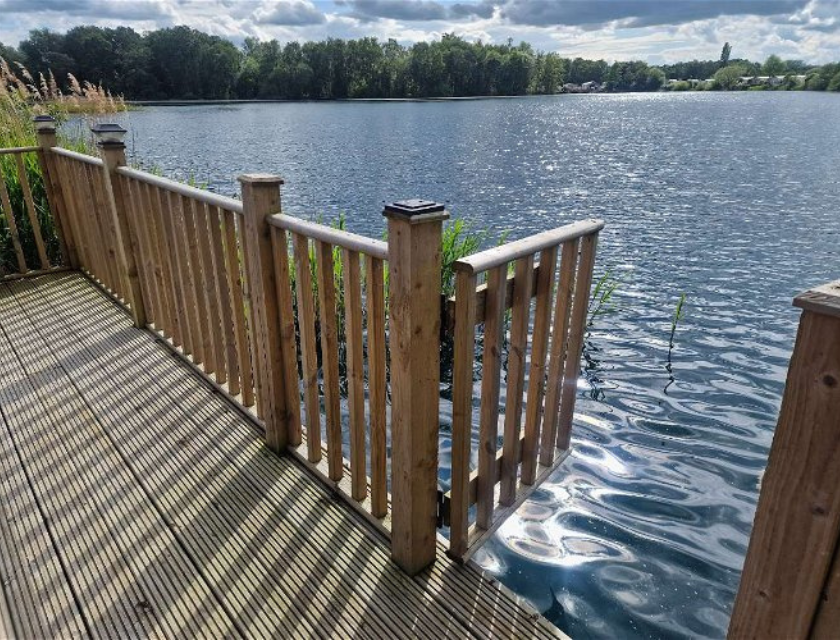 A wooden deck with vertical railings extends out over a body of water. The deck has a gate that opens towards the water, which is calm and reflective. Tall grasses and reeds grow near the water's edge on the left side of the image. In the background, there is a dense line of trees along the far shore under a partly cloudy sky. The sunlight creates sparkling reflections on the water's surface.