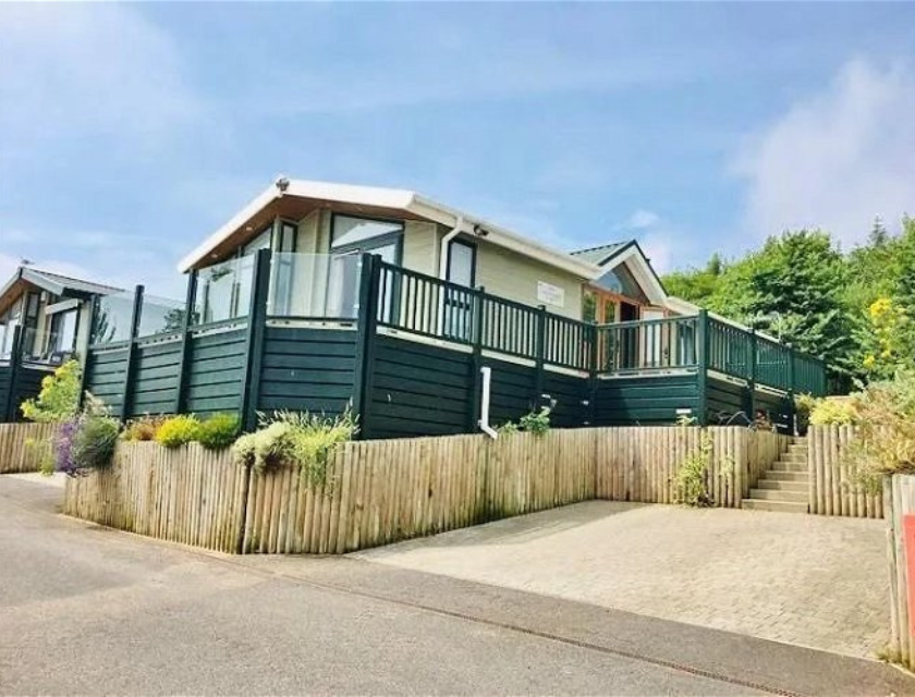 Exterior view of a modern, elevated caravan with a spacious deck surrounded by well-maintained landscaping and wooden fencing. The caravan is situated on a paved driveway with stairs leading up to the entrance, set against a backdrop of greenery and a clear blue sky.