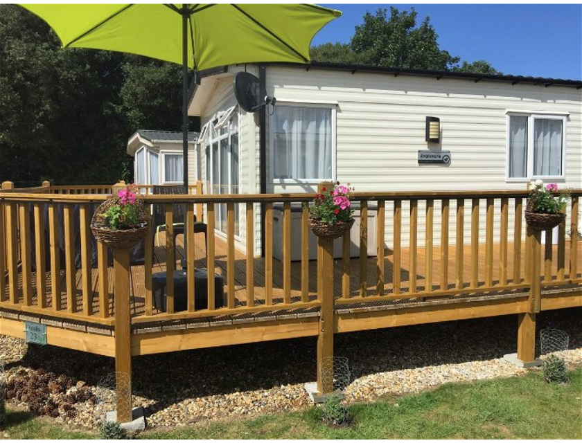 A static caravan located in Pinewoods Caravan Holiday Park in Norfolk, featuring a wooden deck with hanging flower baskets. This cozy retreat is perfect for those looking to explore the scenic walking trails in Norfolk. The park is surrounded by lush greenery, providing easy access to beautiful nature trails.