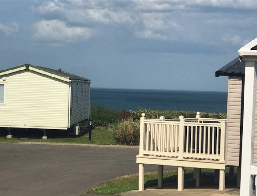 A coastal holiday park with mobile homes overlooking the sea. The foreground features a small wooden deck adjacent to one of the homes, providing a view of the ocean under a clear sky.