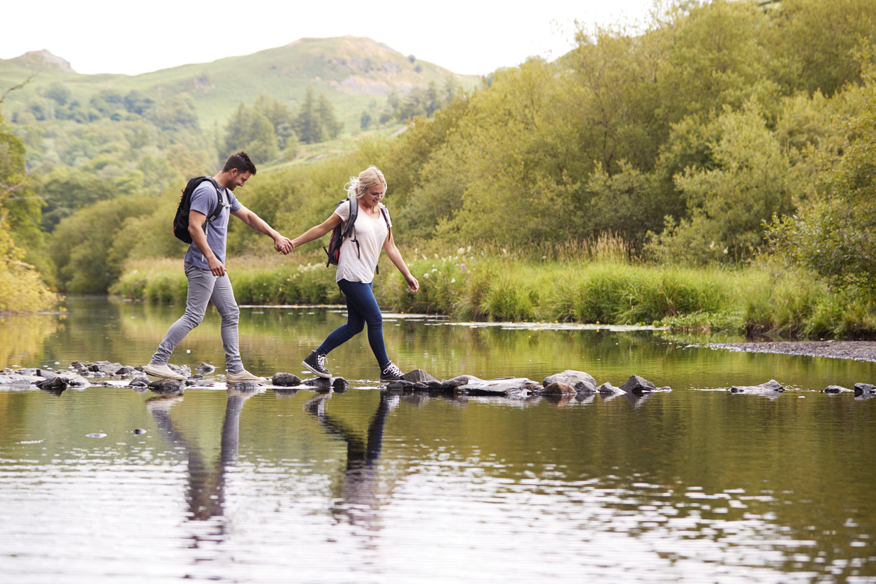 Couple Crossing River Whilst Hiking In UK Lake District