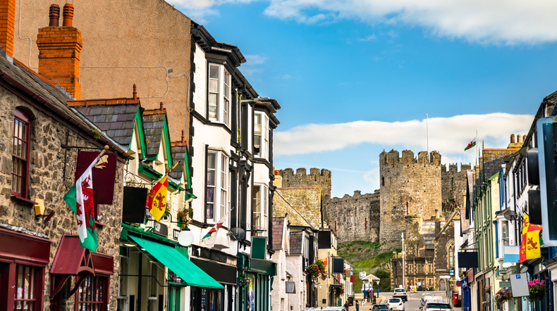 A street in a historic town with colorful shops and buildings, decorated with Welsh flags, leading up to a large stone castle in the background under a partly cloudy sky.