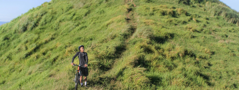 Cyclist riding downhill on a grassy path. The sky in the background is bright blue and cloudless.