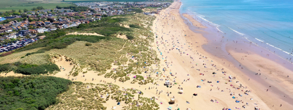 Aerial view of a wide sandy beach with scattered beachgoers and green sand dunes, leading to a coastal town under a clear blue sky.