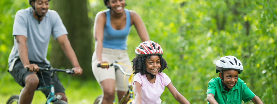 Smiling family of four cycling together on a sunny day, with parents and children wearing helmets and enjoying a ride through a green, leafy park.