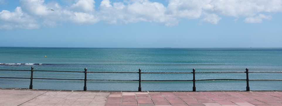 A seaside promenade with a black railing, overlooking calm turquoise ocean waters under a bright blue sky with scattered clouds.