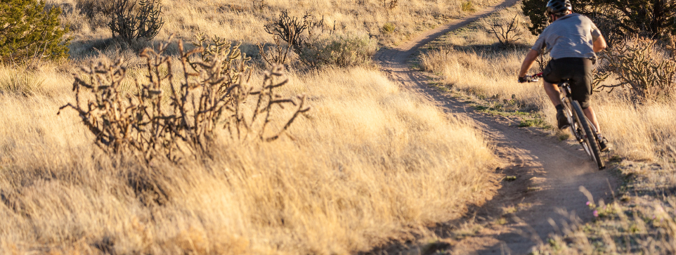 Single rider turning a corner on a bike on a cycling trail. The trail is dusty and the picture is taken with a sunrise/set in the background. 