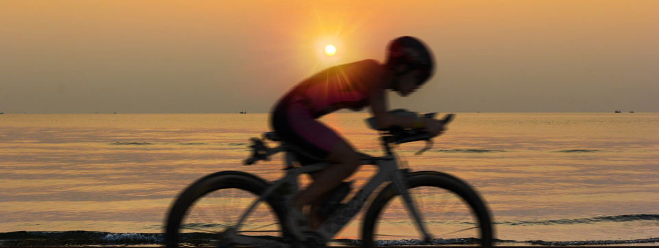 Challenging cycling trails can be found in Cornwall. A single rider with their head down while cycling. The background shows a sunser over pristine waters. 