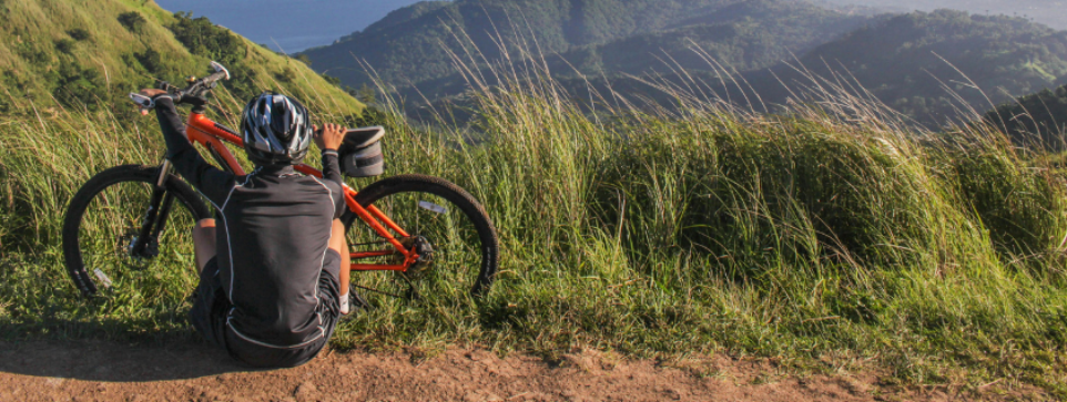 Male cyclist resting by bike, overlooking a vast mountainous landscape. The gras in the foreground is blown by the wind. The background shows mountains and the sky is bright blue with a single cloud visible.