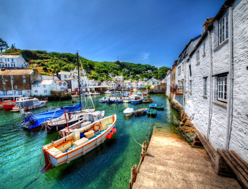 A picturesque coastal village with colorful boats moored in a clear, tranquil harbor, surrounded by whitewashed buildings and lush green hills under a bright blue sky.