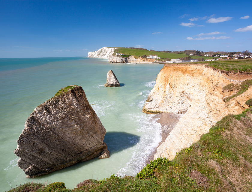 Coastal cliffs & sea stacks overlooking turquoise waters, white chalk cliffs & a seaside village visible in the distance under a bright, clear sky.