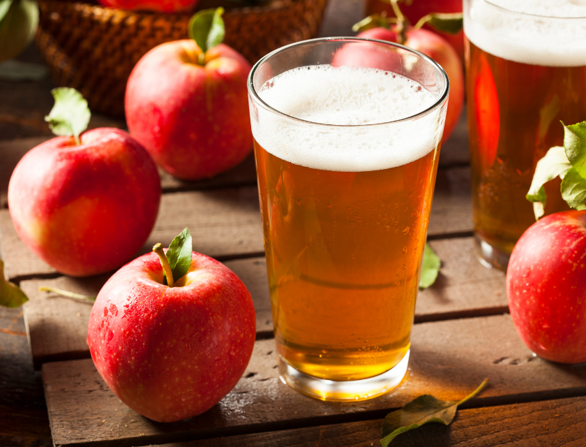 Close up of a wooden table with a pint of apple cider and three red apples visible.