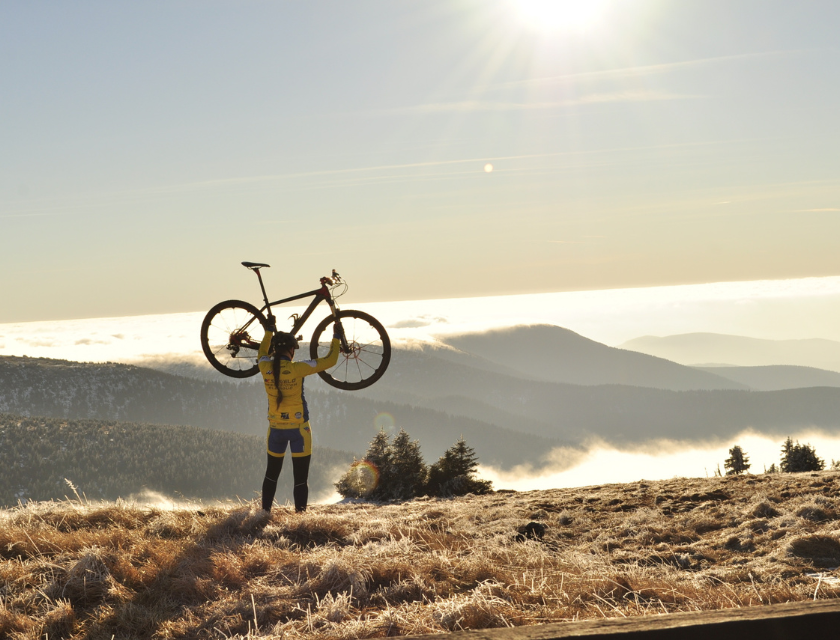 A cyclist in a yellow outfit lifts their bike triumphantly above their head while standing on a frosty hilltop, with the sun shining brightly and a sea of clouds covering the valley below, symbolizing the exhilaration and achievement of reaching the summit on a cycling journey.