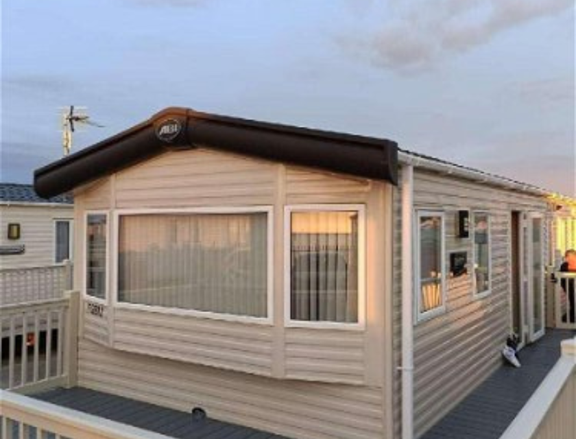 Cream-coloured static caravan with white windows and a dark brown roof trim, set on a raised deck with white railings. The photo captures a warm sunset glow, with blinds drawn inside and a roof aerial visible. Located in a UK holiday park.