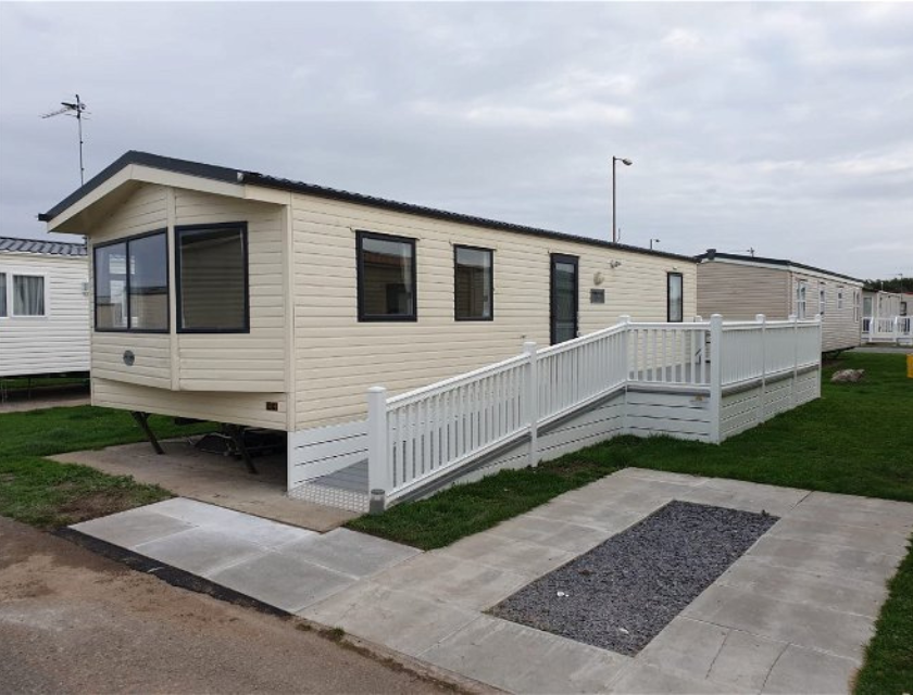 Beige static caravan with a dark grey roof and black window frames, featuring a white ramp for wheelchair access. Located on a concrete base with a gravel area and surrounded by green grass in a UK holiday park.