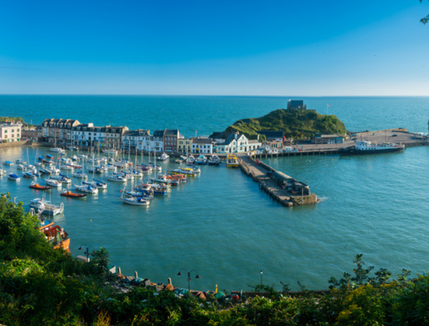 Scenic view of a marina in Devon, England, with boats docked in a harbor surrounded by charming buildings, against a backdrop of clear blue skies and the expansive sea.