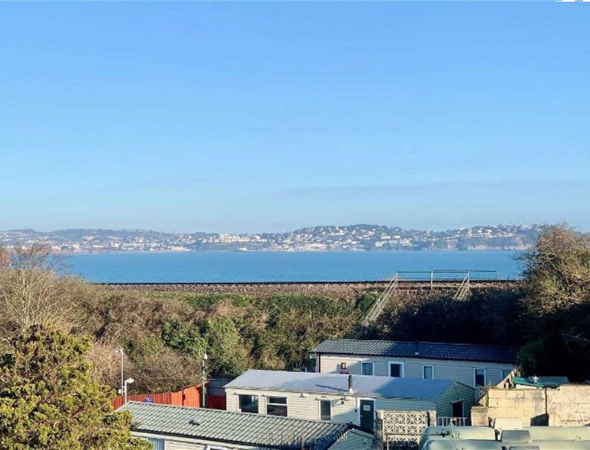 View of a coastal town across the bay in Devon, England, seen from a hillside overlooking holiday homes, with the sea and clear blue sky in the background. A great spot for those searching for things to do in Devon, offering scenic views and peaceful surroundings.