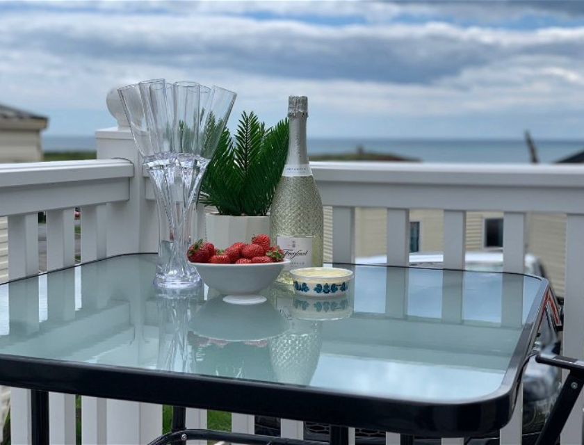 A glass table set on a deck overlooking the ocean is arranged with a bottle of sparkling wine, a bowl of fresh strawberries, and a small potted plant The setting evokes a sense of relaxation and enjoyment, perfect for a peaceful moment by the seaside in Devon