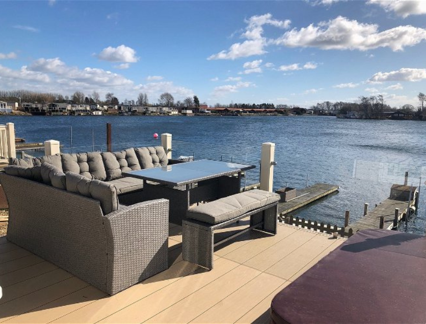 An outdoor seating area at Tattershall Lakes in Lincolnshire with cushioned furniture and a table is set on a deck overlooking a peaceful lake, offering a serene spot to relax and enjoy the view on a clear day.