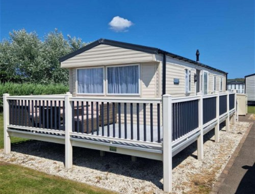 A cream-colored holiday caravan with a raised deck area, surrounded by a well-maintained lawn under a bright blue sky.