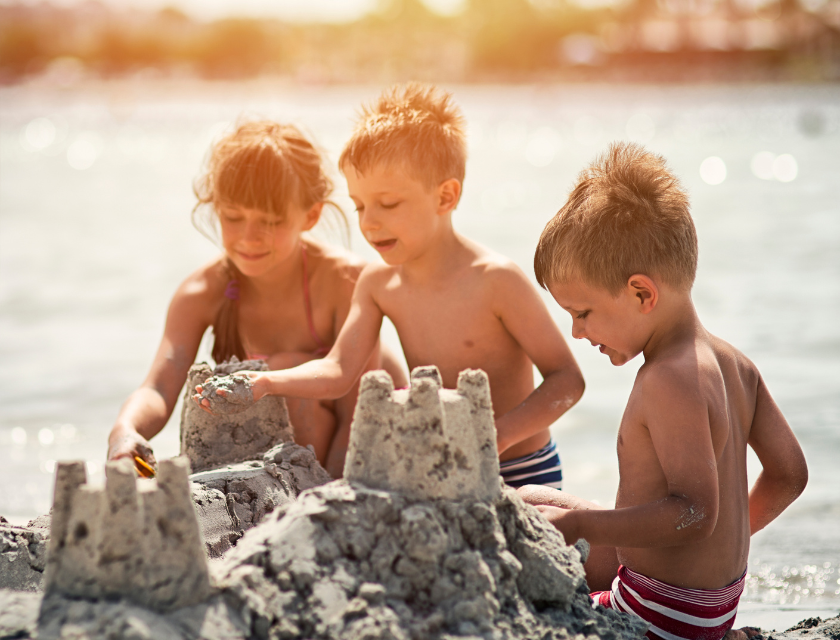 Three young children building sandcastles together on the beach, smiling and enjoying a sunny day by the water.