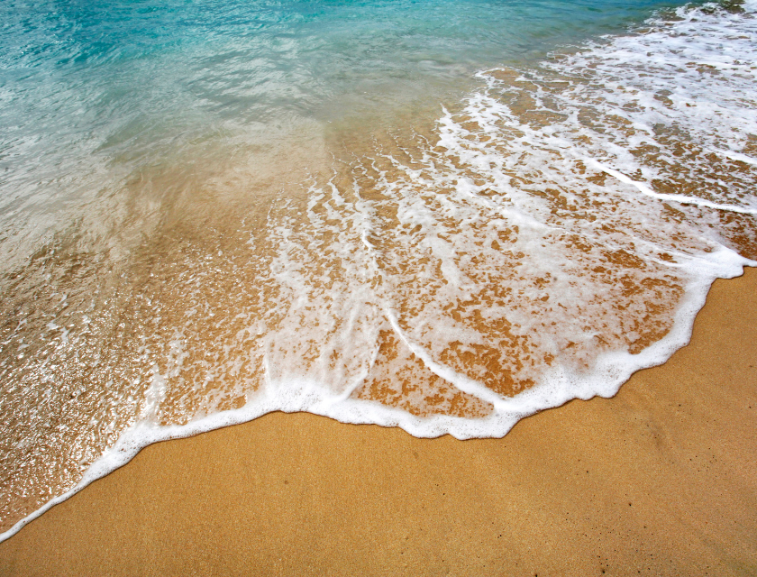 Close-up of waves washing onto a sandy beach, with clear blue water meeting golden sand under a bright sky.