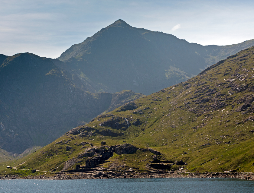 Mountain rising above green hills, with a calm lake at the base and a clear blue sky overhead.