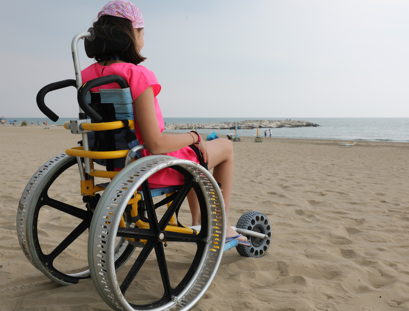 A person sitting in a specialised wheelchair on the sandy beach, looking out at the calm sea. The horizon stretches into the distance.