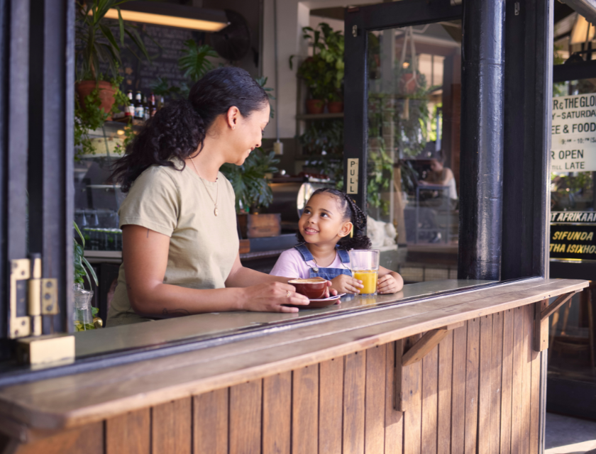 Mother and young daughter smiling at each other while enjoying drinks at an outdoor cafe.