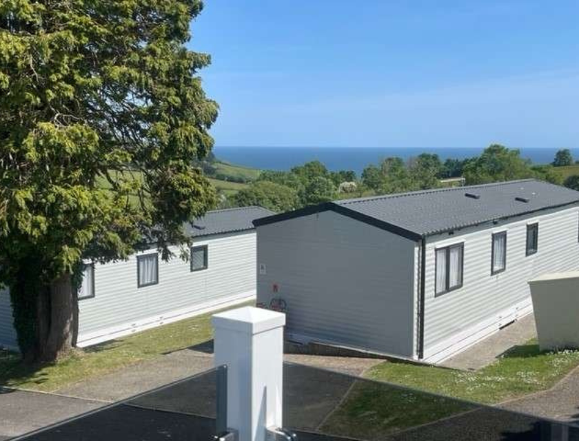 View of light grey static caravans with black roofs in a UK holiday park. The caravans are positioned on a sloped site with a large tree in the foreground. In the background, the image captures a stunning view of the sea and rolling green hills under a clear blue sky.