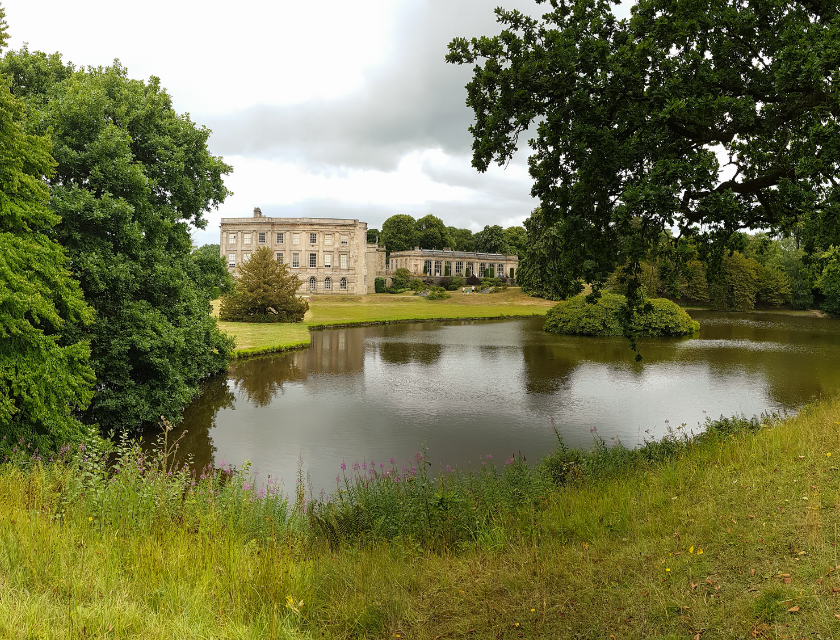 The image depicts a large historic building situated on the far side of a reflective pond, surrounded by lush green trees and well-maintained lawns. The building appears to be a stately home or mansion. The sky above is cloudy and the scene is serene.