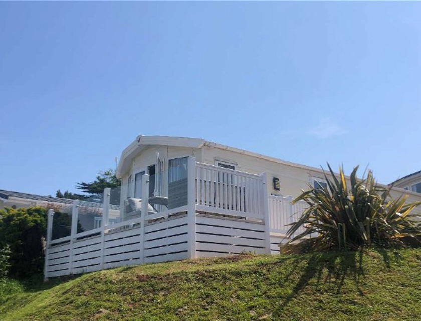 A modern static caravan with a white wooden deck, on a grassy hill under a clear blue sky. The caravan features large windows, a glass door, and outdoor seating, surrounded by well-maintained greenery and shrubs.