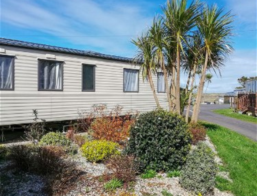 Eternal view of a static caravan at Liskey Hill caravan park near Truro. The sky is bright, blue and clear. The foreground shows well kept trees and bushes, adding an exotic feeling to the mage.