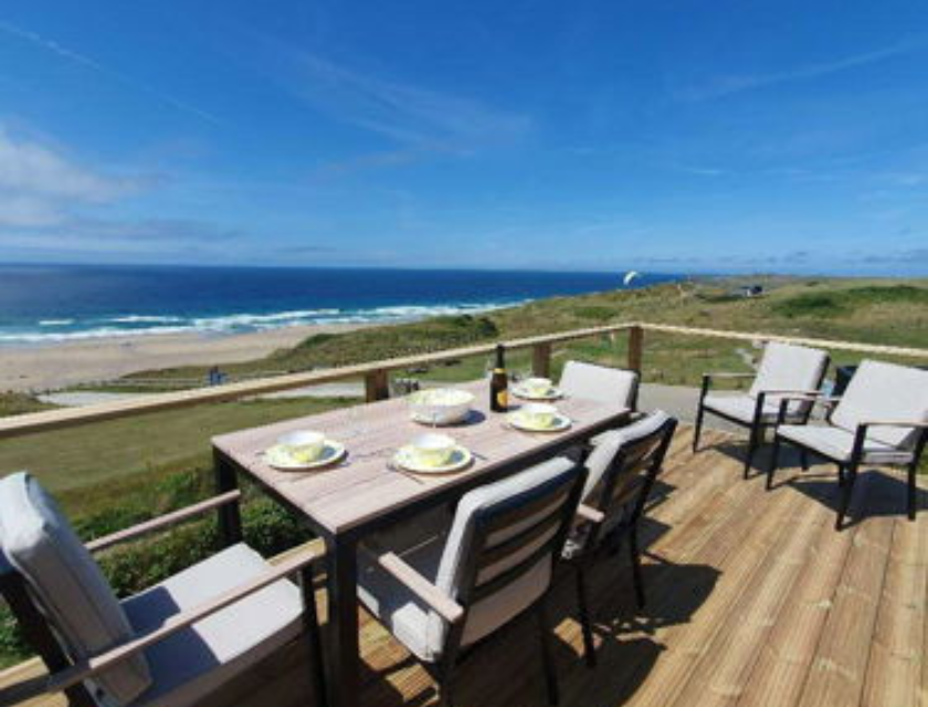 View from the deck of a static caravan at Perran Sands caravan park near Truro. Four chairs are set around a table with four plate places. The day is sunny with bright blue, clear skies. Two lounger chairs are visible in the background.